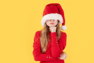 Girl wearing a Santa hat that covers her eyes, with a thoughtful pose on a yellow background. Festive mystery and holiday fun.