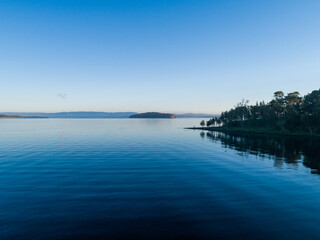 Trees on shoreline of lake with water reflecting blue sky on cold morning seen from aerial view
