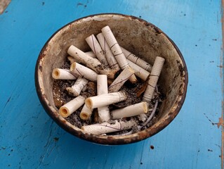 Old dirty metal ashtray filled with cigarette butts on blue wooden background.