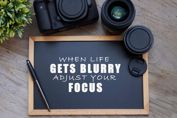 View from above of a black chalkboard with motivational quote saying "When Life Gets Blurry, Adjust Your Focus". Lens, a camera are placed next to the chalkboard on the wooden table.