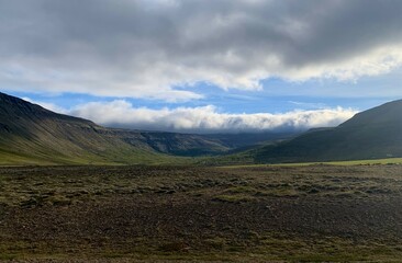 Fototapeta premium A ast valley with rugged mountains and low-hanging clouds, creating depth and isolation. Sunlight highlights the contours of the landscape, emphasizing Iceland's remote, serene beauty.