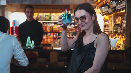 Beautiful woman guest zipping to taste in Christmas blue Lagoon cocktail with couple sitting on luxurious counter bar green neon snow Christmas pine tree festive theme on wine background. Vinosity.