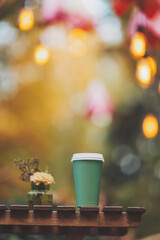 A cup of coffee on wooden table in garden. Coffee in a disposable dish on the background of lights bokeh. Small cardboard blank takeaway coffee cup on table in park. Close-up. Hot drink