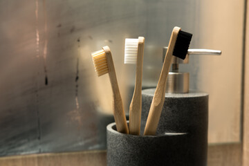 Eco-friendly bamboo toothbrushes with beige, white, and black bristles in a gray holder. Background features a reflection in a mirror and a metallic soap dispenser. Ideal for sustainable living themes