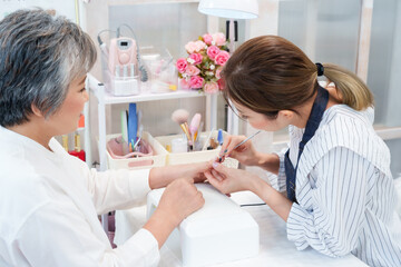 Obraz premium Young female nail technician meticulously filing nails of elderly Asian client in bright, organized nail salon. Technician's focused attention and workspace with various nail tools create of care