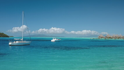 Two sailboats yachts anchored in calm turquoise water of tropical lagoon with overwater bungalows....