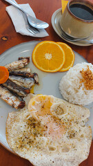 Overhead view of fried egg, sardines, rice topped with garlic, orange slices, coffee cup on a wooden table showing an upgrade on the traditional Filipino breakfast meal
