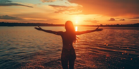 Fototapeta premium A silhouetted woman stands in the water at sunset, arms wide open, soaking in the tranquility of nature.