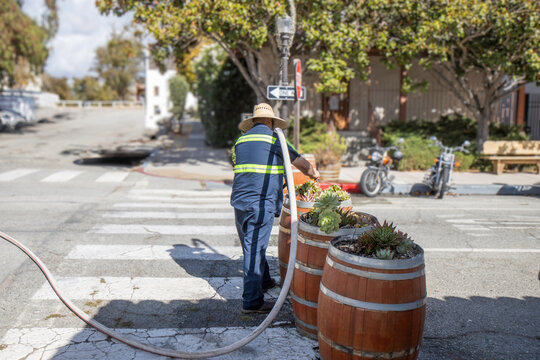 A municipal worker wearing work gear and a straw hat watering planter barrels outside from a hose
