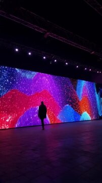 Silhouetted figure walking past a massive LED display screen with vibrant, abstract colors, creating a mesmerizing digital art experience.