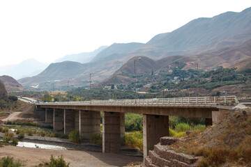 Naklejka premium The way of the bridge over the river with meadows and mountains and cloudy sky