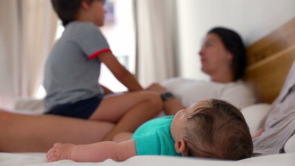 Fototapeta premium Baby lying on a bed with a young boy and mother in the background, enjoying a playful family moment in a relaxed home environment