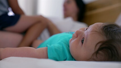 Close-up of a baby lying on a bed while the mother and young boy interact in the background, capturing a peaceful and relaxed family moment at home