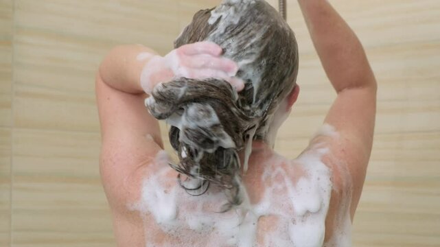 Woman washing her hair, rinsing off foamy shampoo under shower while standing in bathroom, back view. Hair care concept.
