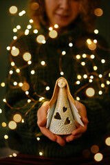 Close-up of hands holding angel figurine with Christmas bokeh background. Woman hands gently holding glowing ceramic angel, concept Christmas and Angel Day spirit in cozy atmosphere with bokeh lights