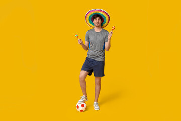 Young male football player with sombrero, soccer ball and maracas on yellow background