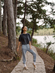 Naklejka premium Young woman in casual outfit standing on a forest pathway, enjoying nature by the lake with a serene expression Outdoor lifestyle and connection with nature concept