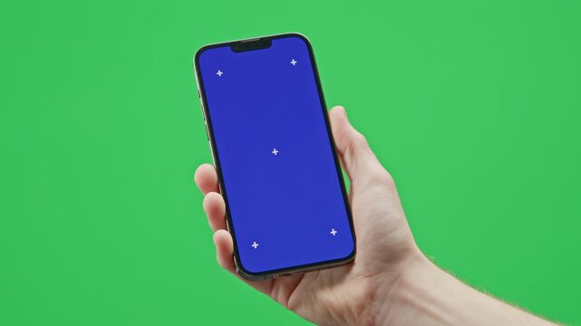 A modern smartphone is held in a man&rsquo;s hand against a green background. The phone of the screen has a chromakey blue image with tracking markers.