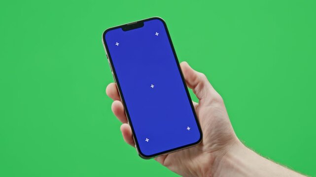 A modern smartphone is held in a man&rsquo;s hand against a green background. The phone of the screen has a chromakey blue image with tracking markers.