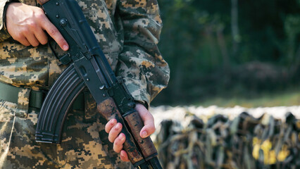 Fototapeta premium Soldier Holding Rifle in Camouflage Uniform Outdoors. A close-up view of a soldier holding a rifle with a sturdy grip, dressed in digital camouflage uniform, standing outdoor.