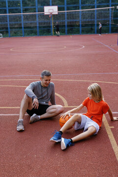 Son bouncing ball on ground sitting on basketball court beside father
