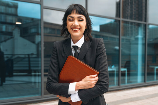 Professional woman in a sleek suit smiles confidently while holding a leather portfolio outside a modern office building