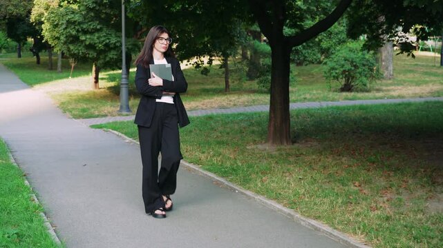 A girl in glasses holds a laptop in her hands while walking through a green park. a woman in a classic black suit looks like a lawyer or an office worker.