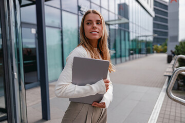 A confident young woman is working with her laptop outside of a modern office building
