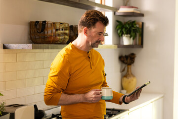 senior man in kitchen holding coffee and reading smartphone, enjoying morning routine, at home