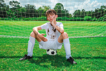 Muddy and sweaty male child youth soccer player sitting on a soccer ball after a game with a disappointed unhappy expression