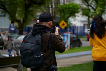 Fototapeta premium Photographer filming with a cell phone to people at a festival in a large man's park with black cap and coffee jacket in black wool lines around cars 