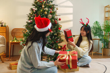 Two young women in Santa hats sitting on the floor, arranging wrapped presents beneath a...