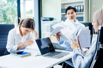 A Muslim businesswoman sits with an Asian man and woman, all middle-aged, at a desk with a laptop, appearing stressed as they discuss challenging business matters in a corporate setting