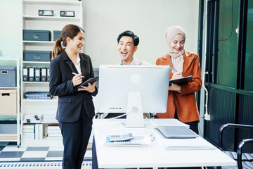 A Muslim businesswoman and an Asian woman sit with a middle-aged businessman at a desk with a laptop, discussing their business plan and analyzing strategies while smiling, shared success