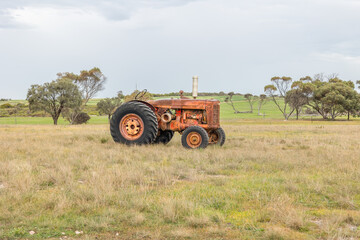 Obraz premium An old orange tractor parked in the middle of a field of grass with some trees on a farm under a cloudy, overcast sky on the Yorke Peninsula in South Australia.