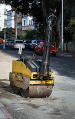 Yellow road roller parked on an urban street, used for compacting soil and asphalt. The roller shows signs of wear and is positioned near an area under roadwork with vehicles visible in the background
