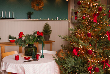 Christmas tree with red ornaments sits in front of a table with a vase of flowers and candles in the dining room