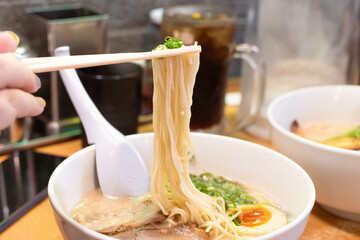 Hand holding chopsticks with yellow noodles of Hakata ramen with sliced pork, boiled egg and green spring onion in white bowl