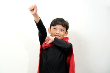 A young boy dressed as a superhero with a red cape, joyfully raising one arm in a heroic pose. Ideal for concepts of imagination, confidence, childhood dreams, and playful energy.