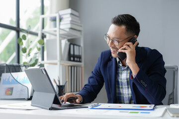 Asian businessman is multitasking, talking on his phone while working on his tablet in his office