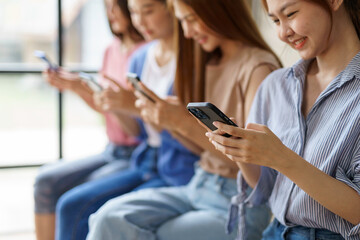 Four young women sitting in a row, engrossed in smartphones, browsing, chatting, and scrolling....