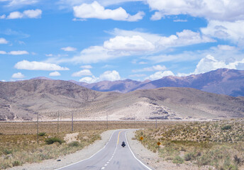 Horizontal shot of a road with a black motorcycle in the distance in a spectacular mountainous landscape at high altitude near the Salinas Grandes in Argentina on a blue sky day with some clouds.
