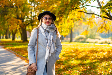 Beautiful middle-aged woman wearing jeans, gray coat, checkered warm scarf, black brimmed hat and sunglasses walking along sidewalk surrounded by big trees in city on sunny autumn day. Front view
