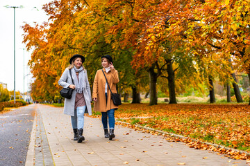 Fototapeta premium Autumn in city. Two young beautiful women wearing coats, jeans and black brimmed hats talking while walking on sidewalk in city on autumn day,golden trees in background. Meeting of friends. Front view
