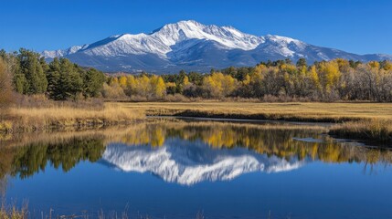 A snow-capped mountain peak reflected in a calm lake with yellow autumn trees in the foreground, under a clear blue sky.