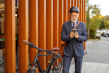 Confident senior businessman using mobile applications while standing with bicycle by wooden wall in city