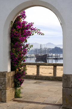 Museum caravel La Pinta in the fishing port of Baiona seen through an arch with purple flowers. Pontevedra, Galicia, Spain.