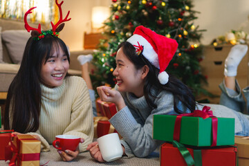  Two friends lying on the floor near a decorated Christmas tree, laughing and enjoying hot drinks,...