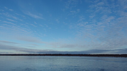 a serene landscape with a large body of water in the foreground. The water is calm and reflects the sky above. The sky is mostly clear with a few scattered clouds, and it has a gradient of blue shades