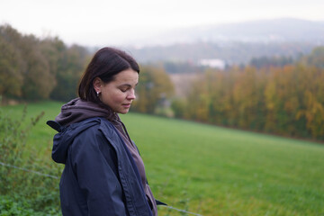 Girl dressed in jacket standing sideways to camera looking down smiling against blurred natural autumn landscape. Nature travel concept